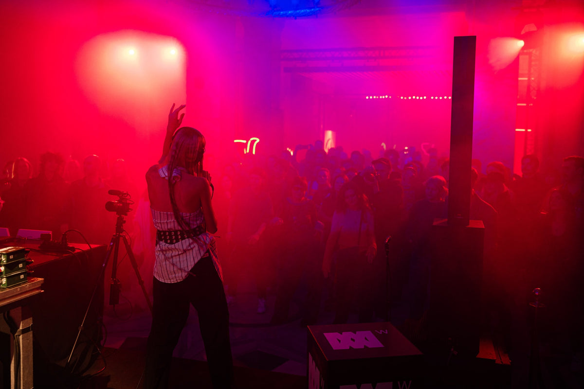 A person stands on a stage in a crowded indoor venue, back turned toward the audience, under heavy red and pink stage lighting with smoke. A crowd is visible in the background, along with speakers and lighting equipment.