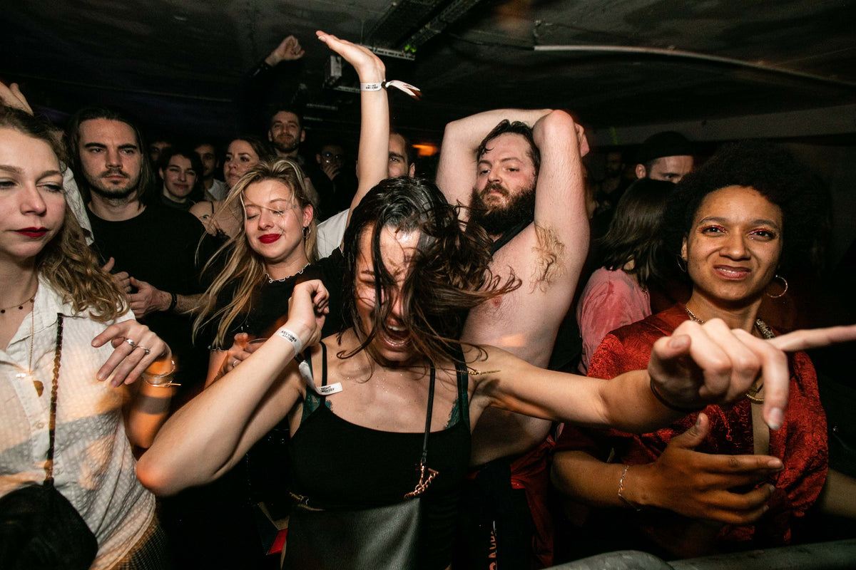A crowded nightclub or party scene with multiple people dancing and socializing in a dimly lit indoor venue. The central focus is a woman in a black tank top dancing energetically, surrounded by other guests.