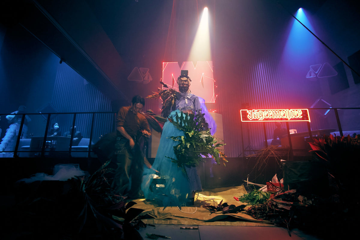 A person in a dramatic blue gown stands on a dark stage under spotlights, holding a large bouquet of flowers, with another person beside them. Neon signs, band equipment, and theatrical lighting create a concert-like environment.