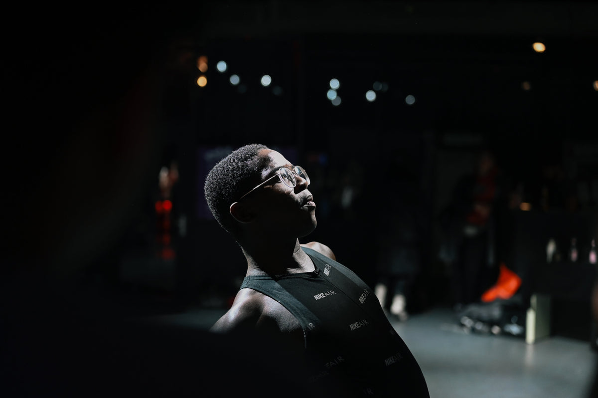 A man wearing a black athletic tank top and glasses, looking upward in a dimly lit gym or studio environment.