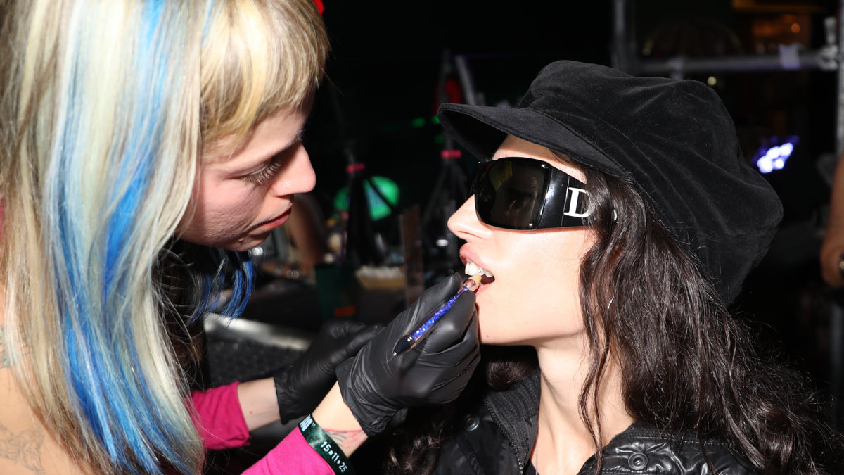 A close-up lifestyle shot of a makeup artist applying lip gloss to a model wearing a black velvet hat, black sunglasses with a silver 'D' logo, and a black jacket. The artist has blonde hair with blue streaks and is wearing black gloves.