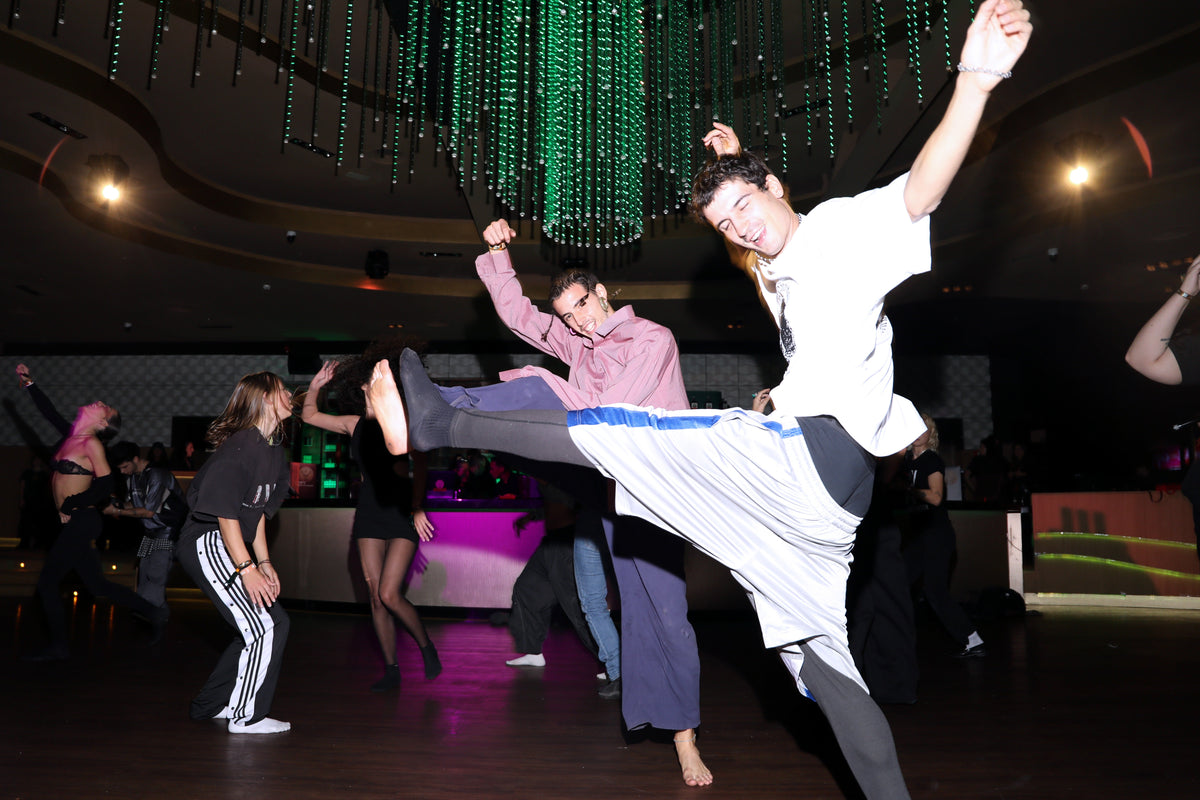 A man in a white t-shirt and gray sweatpants kicks high during an energetic dance party in a dimly lit hall with green hanging lights and other dancers in the background.