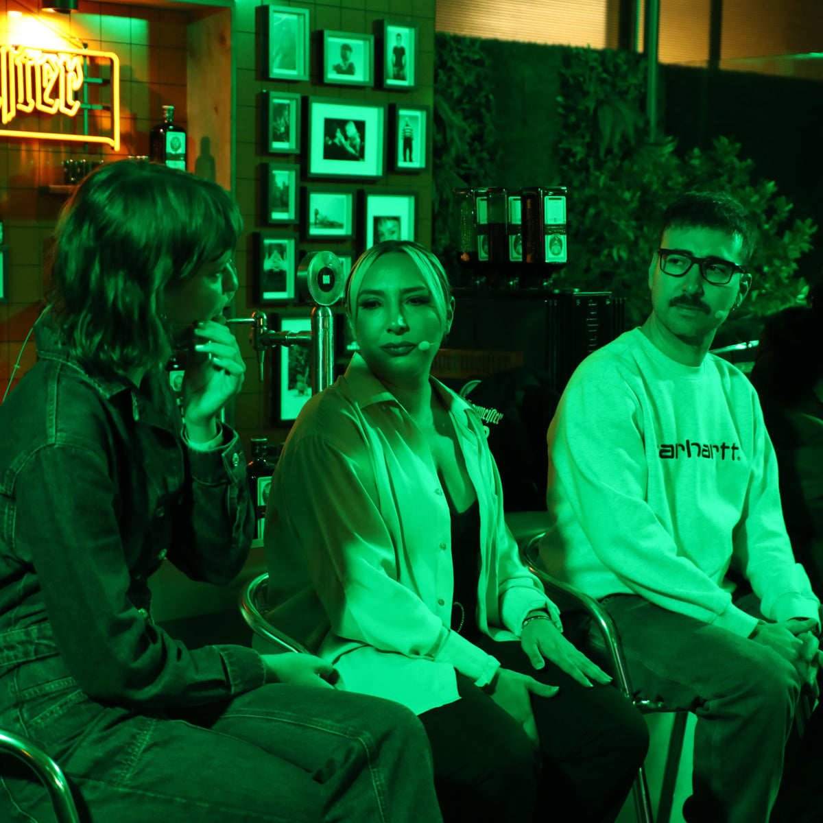 Three people seated indoors under strong green lighting, with framed wall art and neon signs in the background. The person on the right wears a white Carhartt sweatshirt.