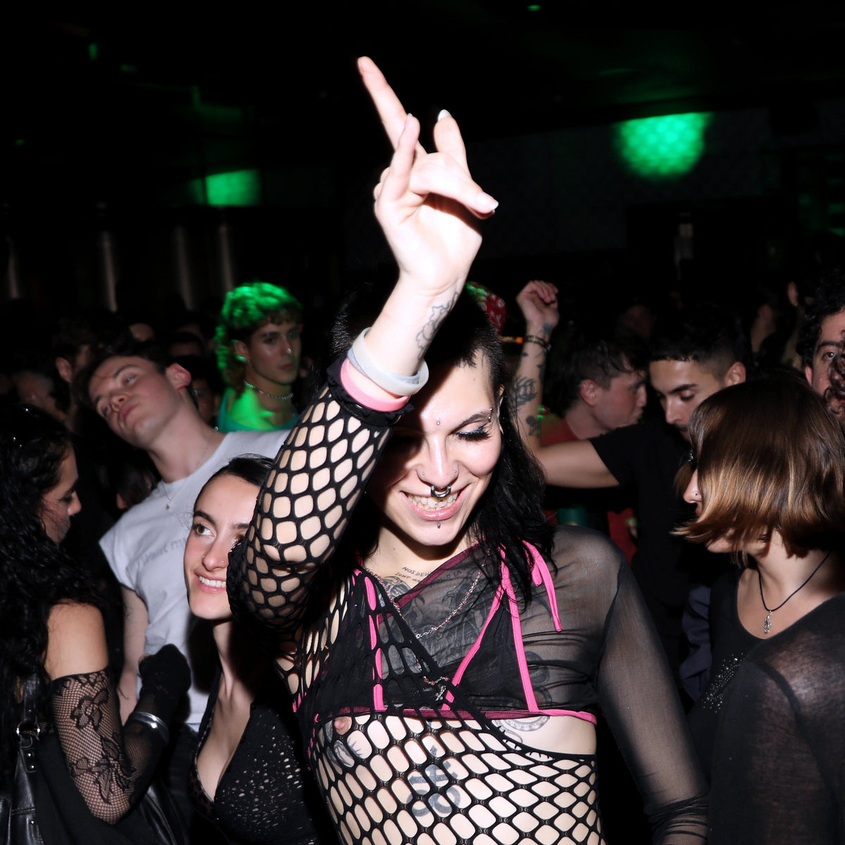 A woman at a nightclub wearing a black fishnet top with pink trim, posing with her hand raised in a gesture while surrounded by a dancing crowd under green party lights.