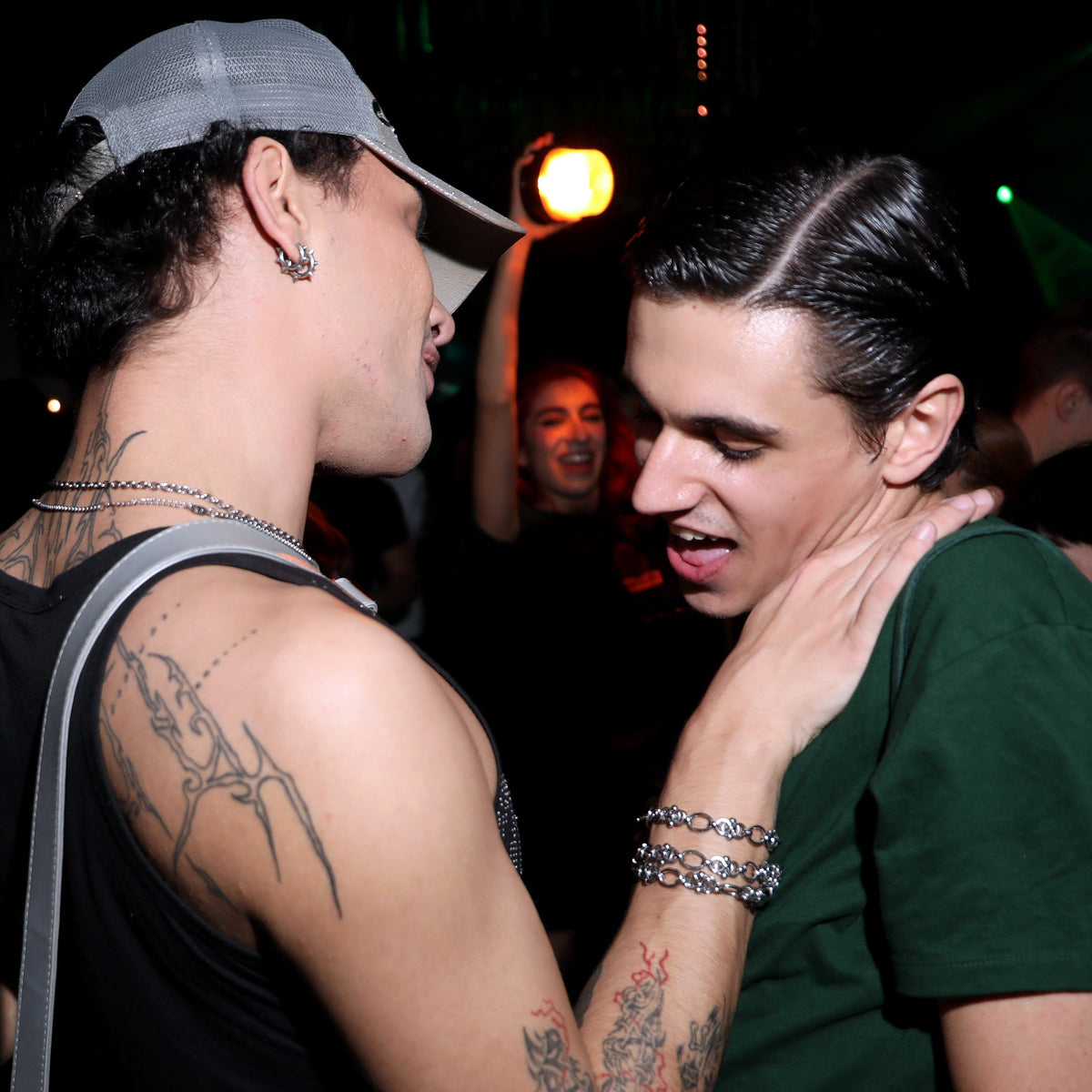 Two young men dancing closely at a nightclub. The man on the left wears a gray baseball cap, a black tank top, and silver chain jewelry.