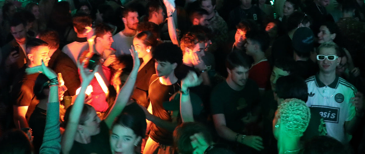 A crowded nightclub or concert scene with multiple people dancing and singing under colored green and orange stage lighting. A person on the right wears a white ASICS track jacket, and another person in the foreground wears 3D sunglasses.