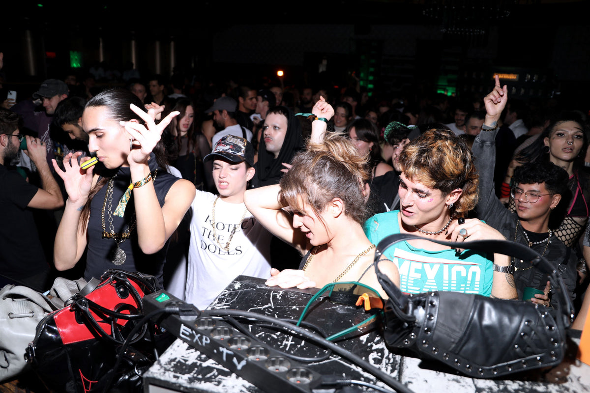 A crowded nightclub or party scene with multiple people dancing and standing around a DJ booth. In the foreground, a black studded handbag and tangled cables sit on the DJ table, with a crowd illuminated by dim party lighting in the background.