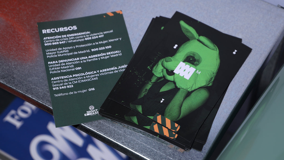 A stack of printed cards or brochures on a metallic surface. One card is a dark green informational sheet titled 'RECURSOS' with white text, and another card features a green rabbit-like character holding a white sign with '100%'.