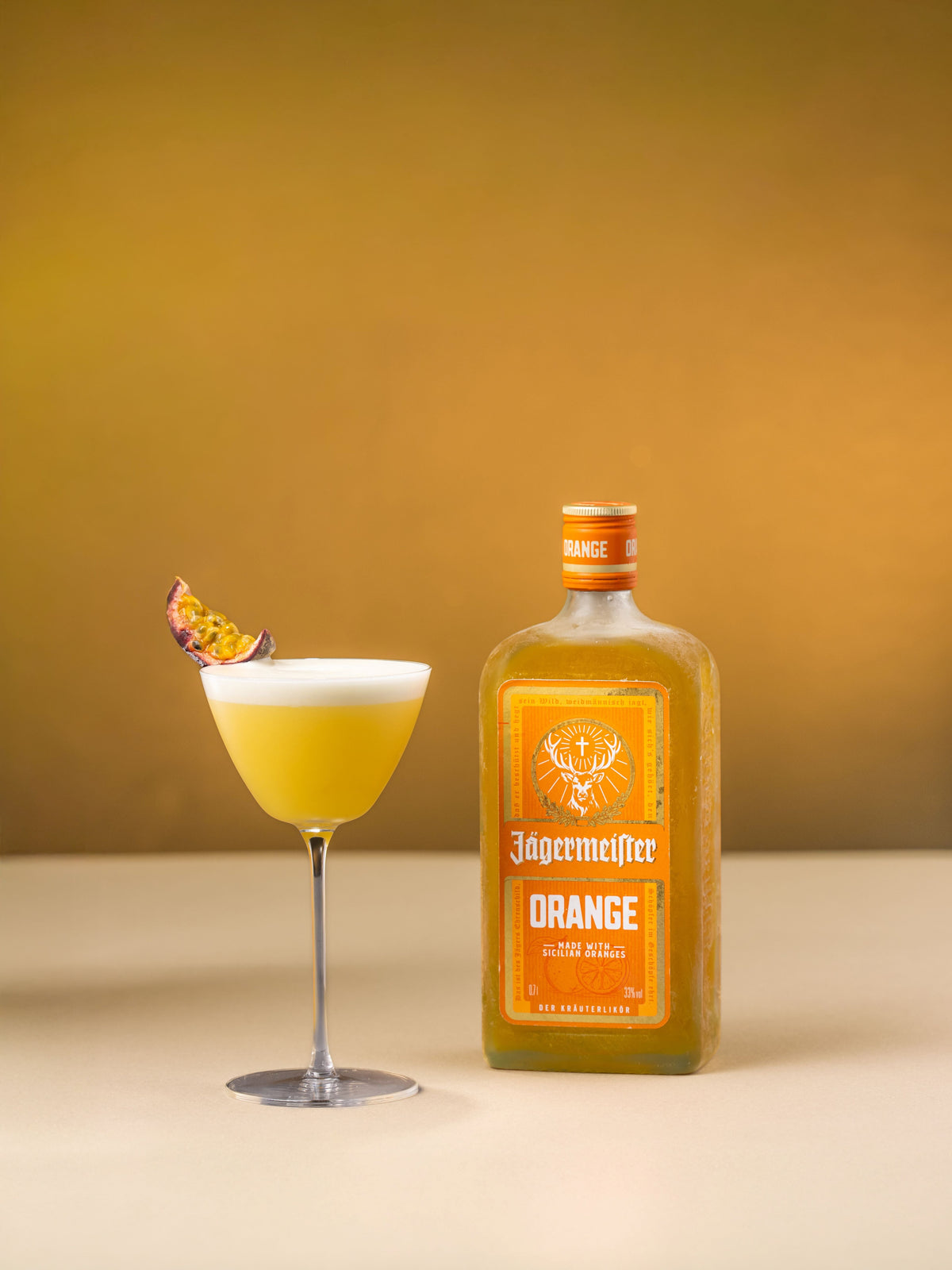 A bottle of Jagermeister Orange liqueur stands next to a coupe glass filled with the same orange-colored drink and garnished with a passion fruit twist, set against a warm golden background.