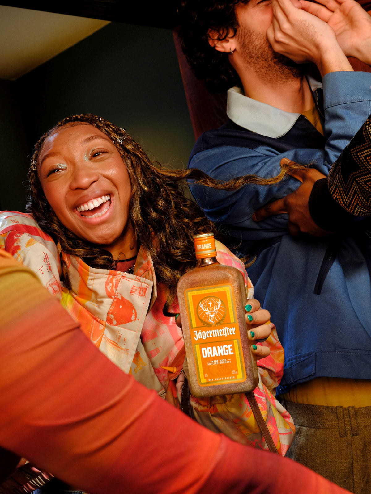 A person in a colorful patterned top holds a bottle of Jägermeister Orange Liqueur in a candid, playful indoor setting with another person in the background.