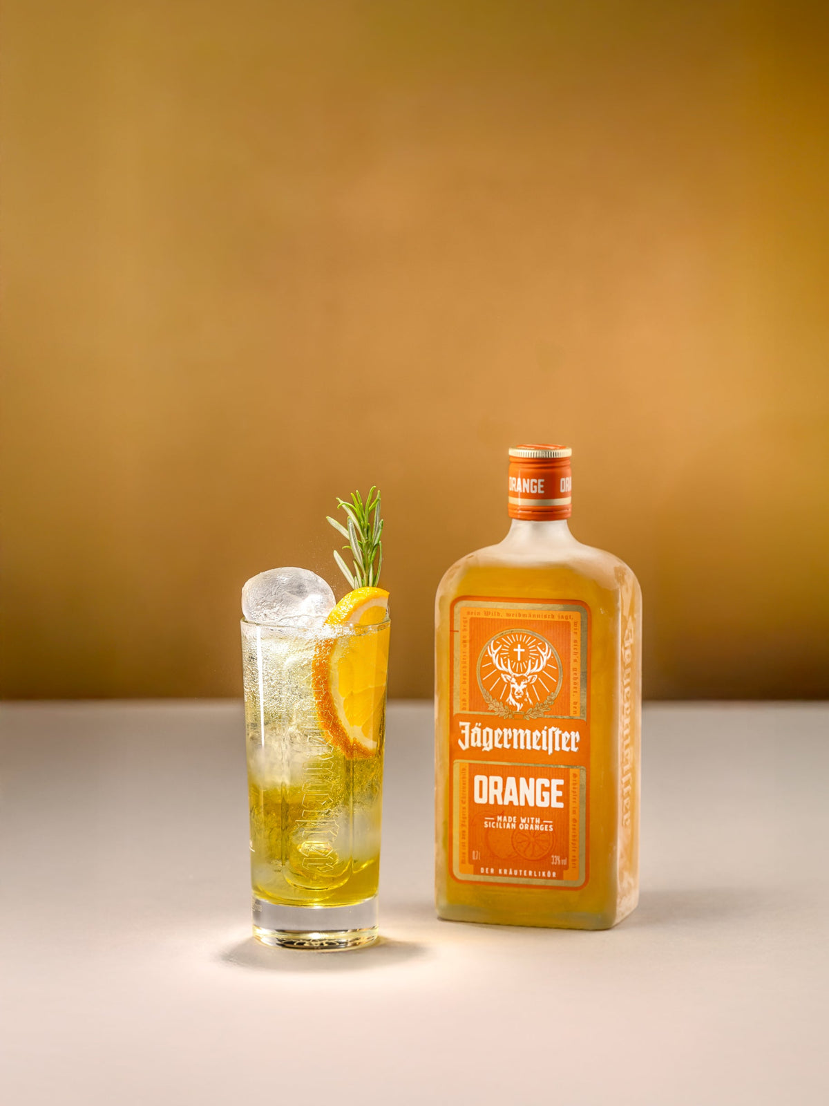 A bottle of Jagermeister Orange alongside a highball glass filled with a golden-orange cocktail, garnished with an orange slice and rosemary, set against a warm gradient background.
