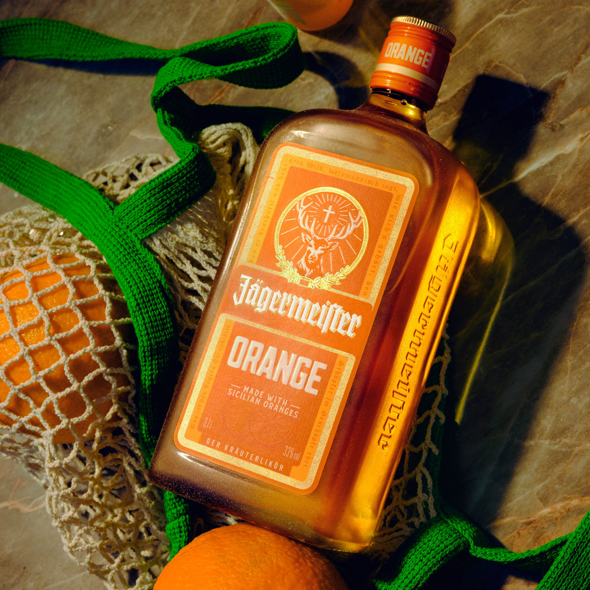 A bottle of Jägermeister Orange liqueur with a prominent orange label, styled on a stone surface with oranges in a mesh bag and green straps around it.