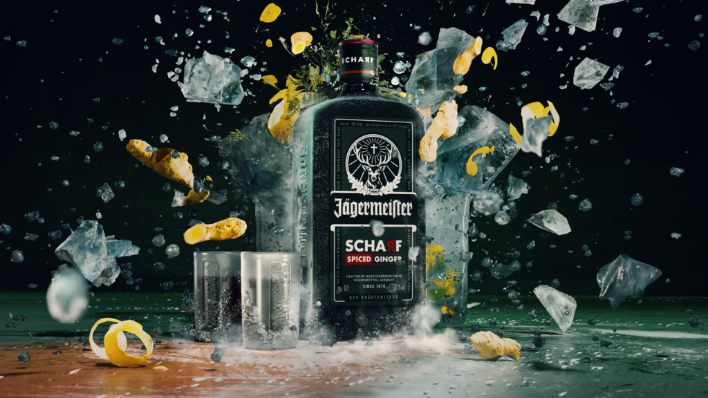 A bottle of Jägermeister Scharf stands in the center of a dramatic splash scene with ice cubes, liquid, and lemon peels flying around it against a dark background.