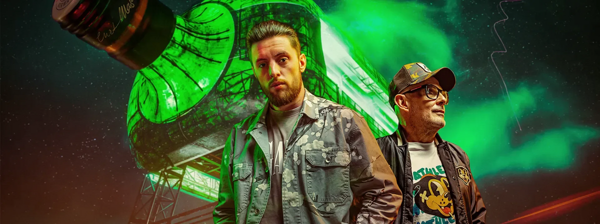 Two men standing in front of the Jägermeister 'Gigant'. The man on the left wears a gray camouflage jacket over a light shirt, and the man on the right wears a dark cap, glasses, and a graphic t-shirt under a green glow.