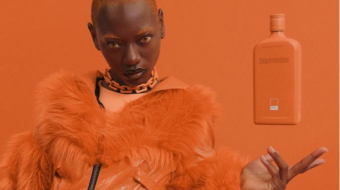 A model in a bright orange fur jacket and gold chain necklace poses against a solid orange background, with a matching orange Jägermeier bottle floating to the right.