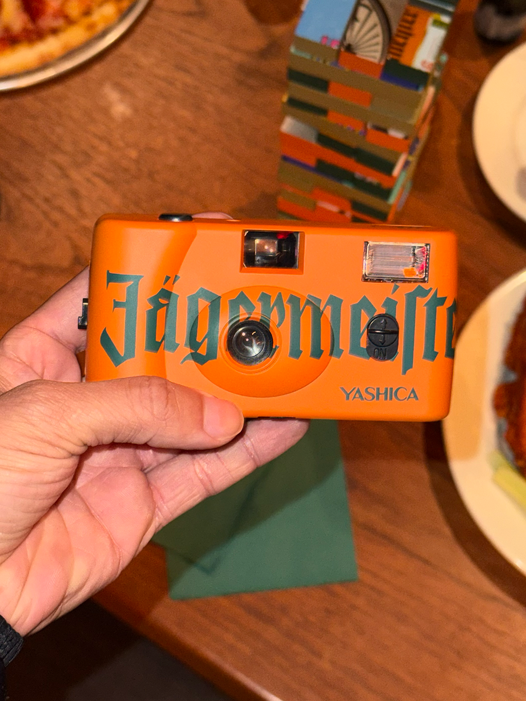 Orange camera with 'Jägermeister' branding held by a hand on a wooden table.