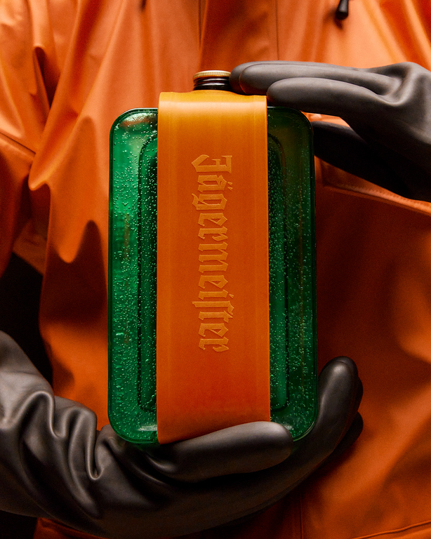 Green and orange cooling device with 'Jägermeister' branding held by a person in black gloves wearing an orange suit