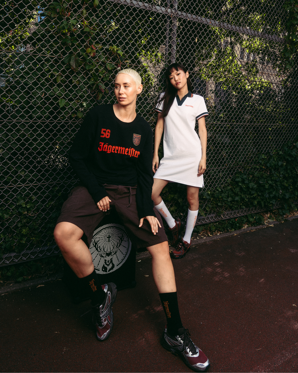 Person wearing a white Jägermeister polo dress standing beside a chain-link fence, with another person seated in front wearing a black Jägermeister sweater and shorts. Outdoor urban setting with greenery in the background.