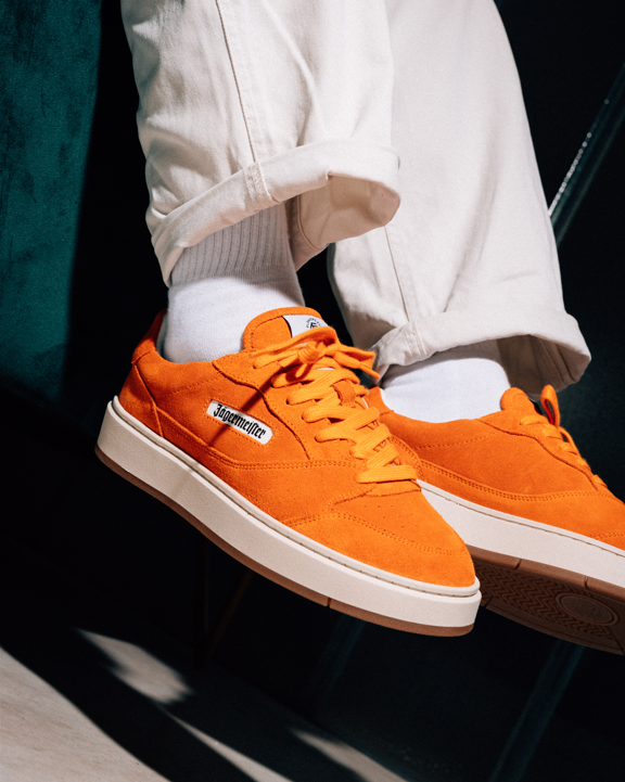 Bright orange Jägermeister sneakers with white soles and white socks, worn with off-white cuffed pants, shown in a styled lifestyle setting.