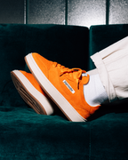Bright orange Jägermeister sneakers with white soles and white socks, worn by a person sitting on a dark teal velvet couch. A white label on the side of the shoe reads 'Jägermeister'.