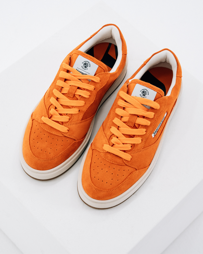 A pair of Jägermeister sneakers in vibrant orange colour, featuring matching orange laces, white midsoles, and white interior lining, photographed on a clean white geometric background.
