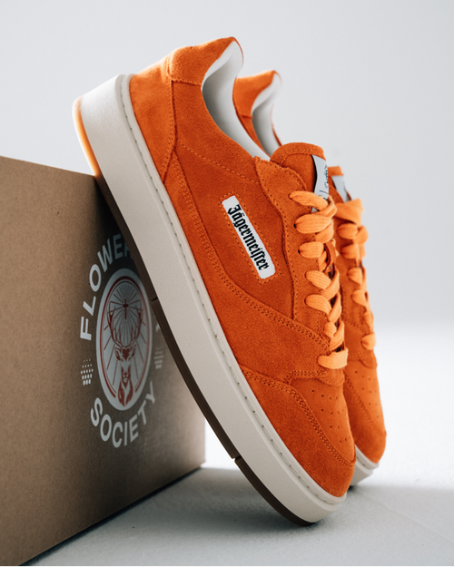 Orange Jägermeister sneakers with white soles and a white side label reading 'Jägermeister', displayed with a brown box printed with 'Flower Society' on a light gray background.