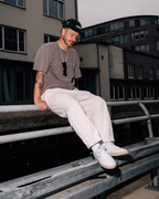 A man wearing a black baseball cap, taupe t-shirt, off-white trousers, and white Jägermeister sneakers sits on a metal guardrail in an urban outdoor setting.