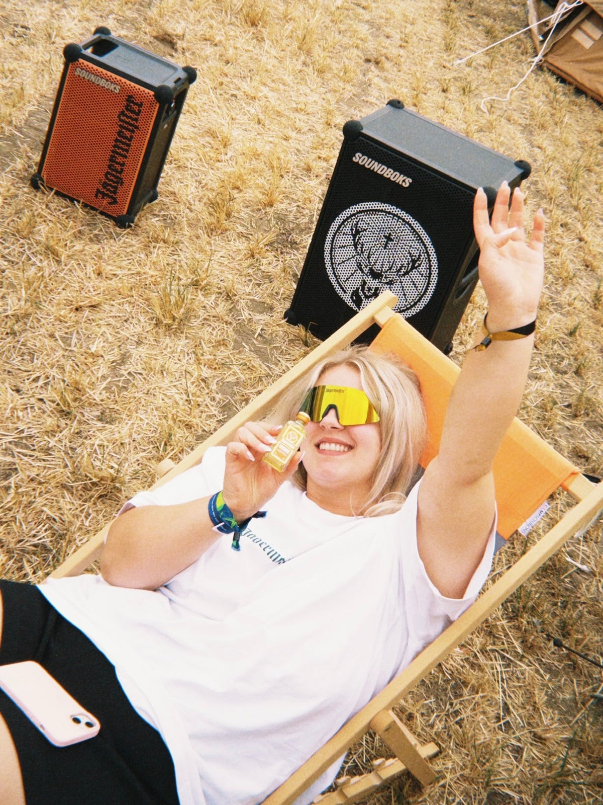 A woman wearing a white t-shirt and yellow-tinted sunglasses relaxes in a wooden lounge chair outdoors on dry grass, with two Soundboks speakers visible behind her.
