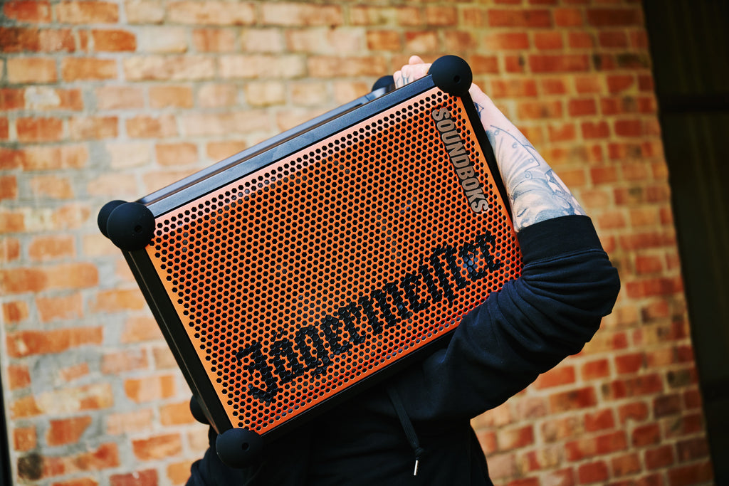 Persoon houdt een draagbare Soundboks-speaker in oranje en zwart met 'Jägermeister'-branding vast voor een bakstenen muur.