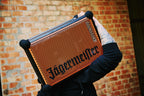 Persoon houdt een draagbare Soundboks-speaker in oranje en zwart met 'Jägermeister'-branding vast voor een bakstenen muur.