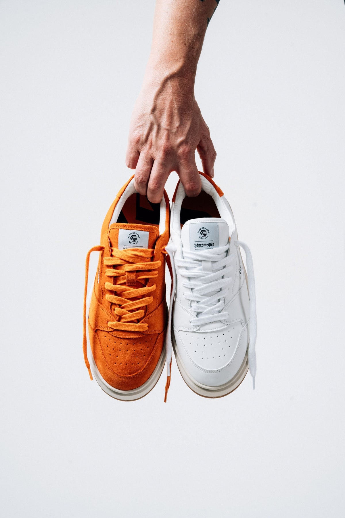 A hand holds up two low-top sneakers against a white background: one bright orange Jägermeister sneaker and one white Jägermeister sneaker, both with matching laces and white soles.
