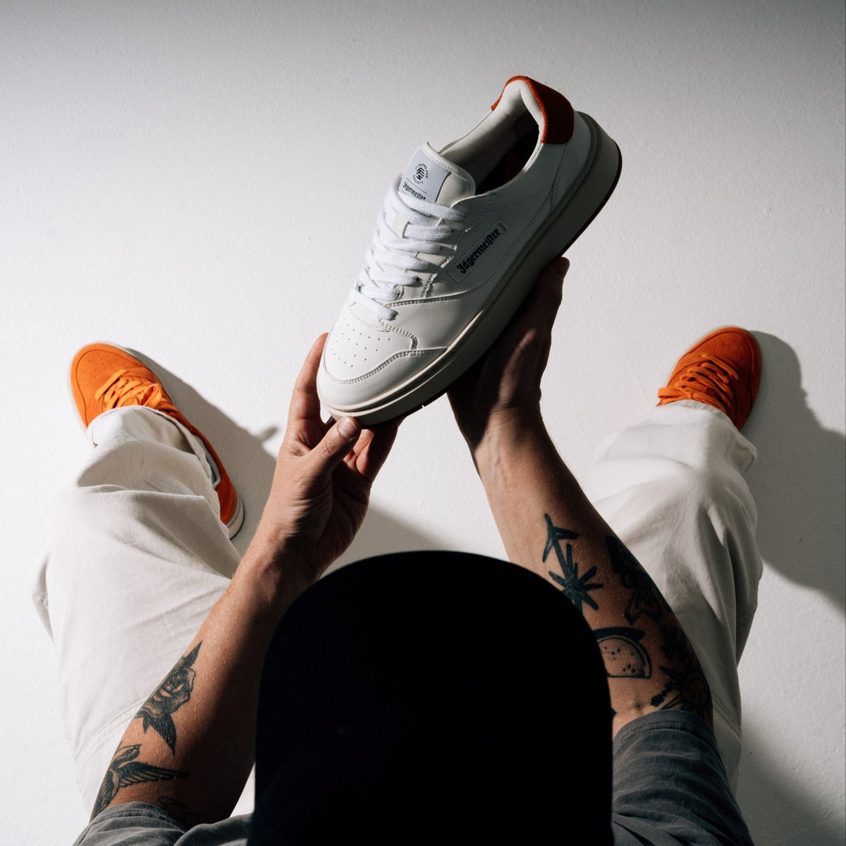 A person wearing white pants and bright orange Jägermeister sneakers holds up a white low-top sneaker with black Jägermeister branding details against a plain white wall.