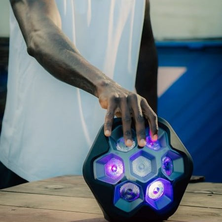 A person in a white shirt holds a black hexagonal speaker with glowing purple and blue LED lights on a wooden surface.