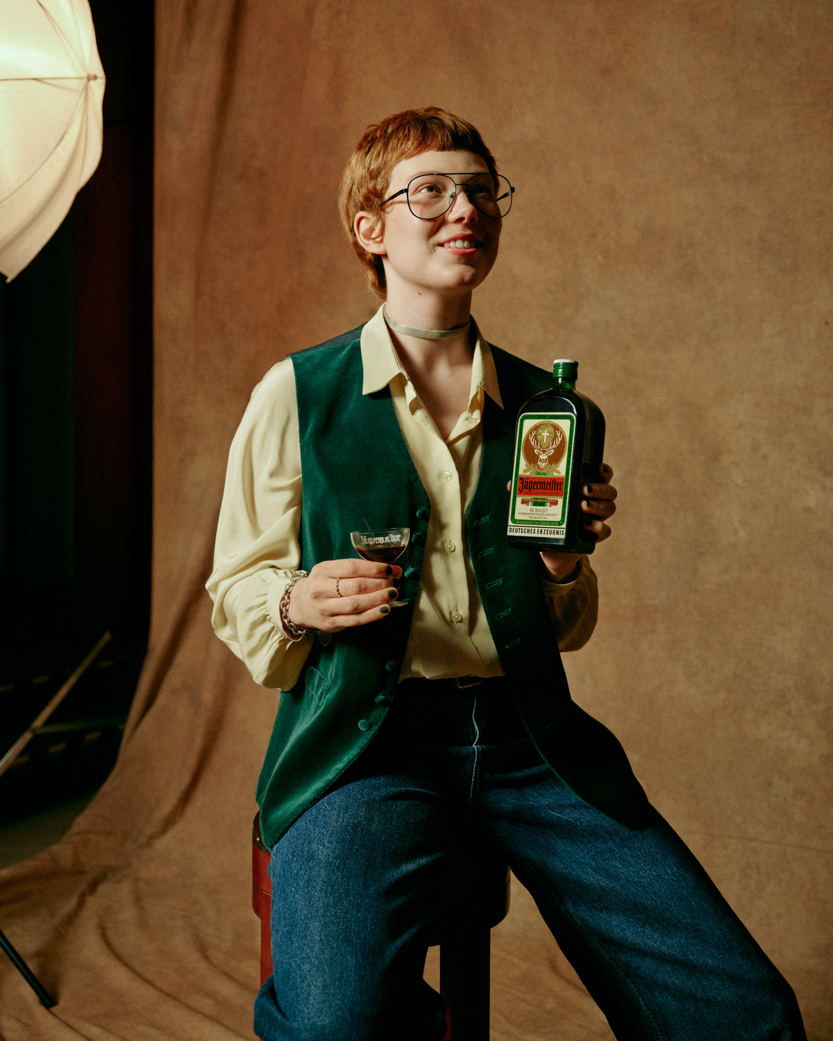 A person with short red hair and glasses holds a retro-bottle of Jägermeister and a small drink in a studio setting with a warm brown backdrop.