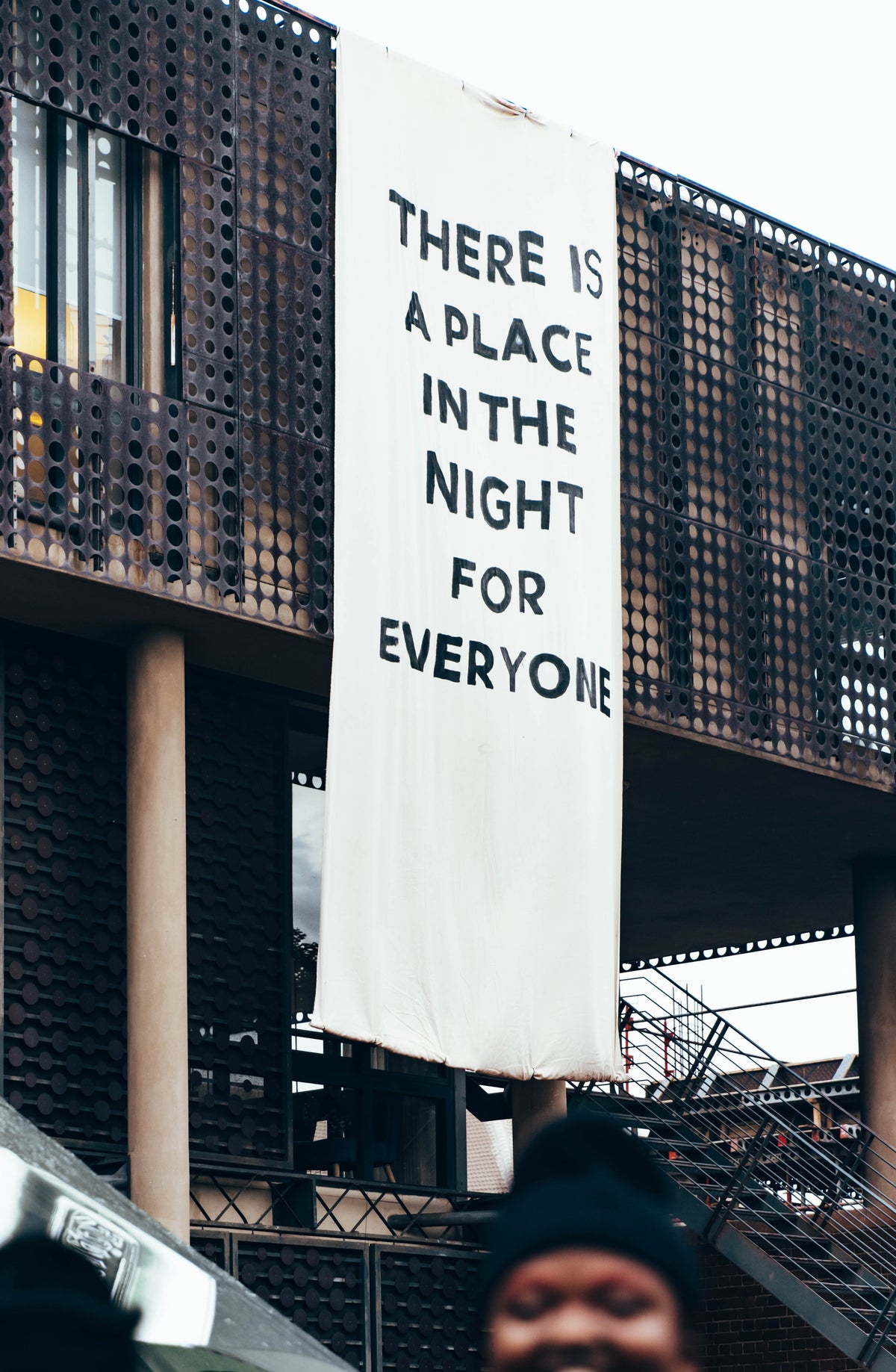 A large white vertical banner hanging from a modern building with a dark perforated metal facade, printed with the text 'THERE IS A PLACE IN THE NIGHT FOR EVERYONE'. The bottom of the banner is slightly worn, and a person in a black beanie is partially visible at the very bottom edge of the frame.