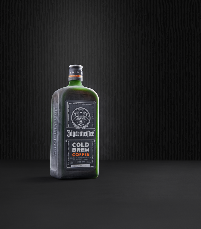 A bottle of Jägermeier Cold Brew Coffee stands on a dark surface against a dark textured background. The bottle is dark green glass with a black label featuring white text and an orange accent.