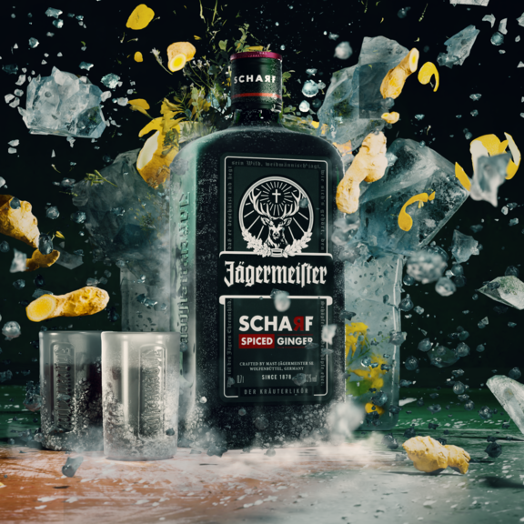 A bottle of Jägermeier Scharf stands in the center of a dramatic splash scene with ice cubes, liquid, and lemon peels flying around it against a dark background.
