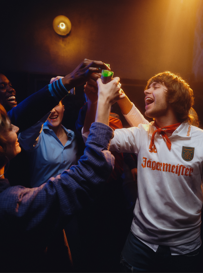 Group of people celebrating with a person wearing a Jägermeister shirt in a dimly lit room.